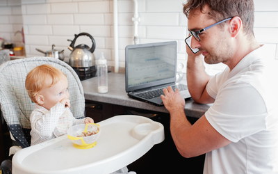 Vater arbeitet am Laptop in der Küche, Kleinkind in Kinderstuhl beim Essen neben sich