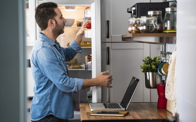 Mann mit Laptop in Küche schaut in Kühlschrank