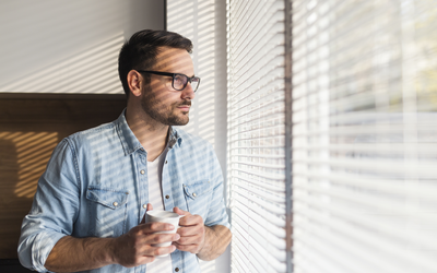 Mann im Homeoffice mit Kaffeetasse schaut sinnend durch Fenster mit Jalousien.