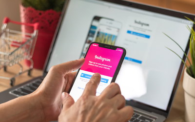 Laptop im Hintergrunde, Hand zeigt auf Smartphone mit Instagram-Seite