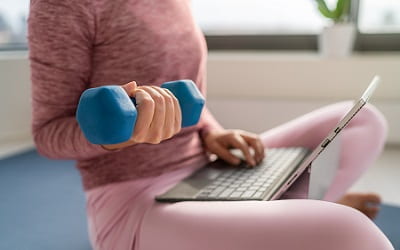 Eine junge Frau sitzt auf dem Sofa, Laptop auf dem Schoß, Hantel in der Hand