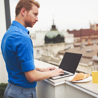 Mann steht mit Laptop auf dem Balkon und schaut hinaus