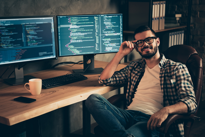Lässig gekleideter jüngerer bärtiger Mann vor Computern schaut zufrieden, IT-Freelancer