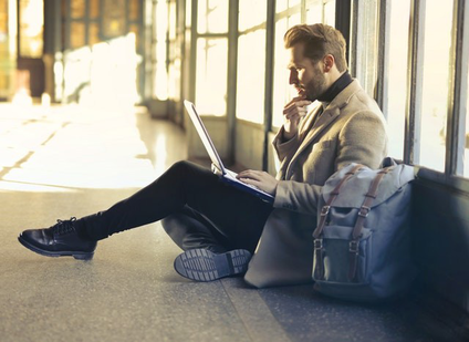 Ein Mann sitzt in einem Gebäude (Flughafen) auf dem Boden und schaut nachdenklich in seinen Laptop