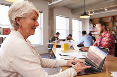 Eine ältere Frau am Laptop im Vordergrund, im Hintergrund jüngere Mitarbeiter in einem Büro
