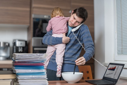 Gestresster Mann im Home Office mit Kind auf dem Arm telefoniert und hat den Laptop im Blick