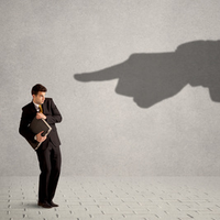 Mann im Anzug mit Aktenordnunger schreckt vor einem Schatten zurück, der mit dem Finger auf ihn zeigt